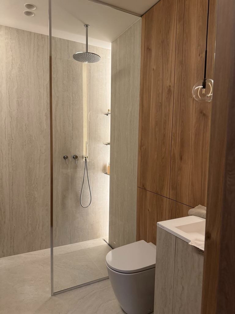 Nowoczesna łazienka z kabiną prysznicową typu walk-in, toaletą i drewnianą zabudową. Minimalistyczny design, jasna kolorystyka, eleganckie oświetlenie.