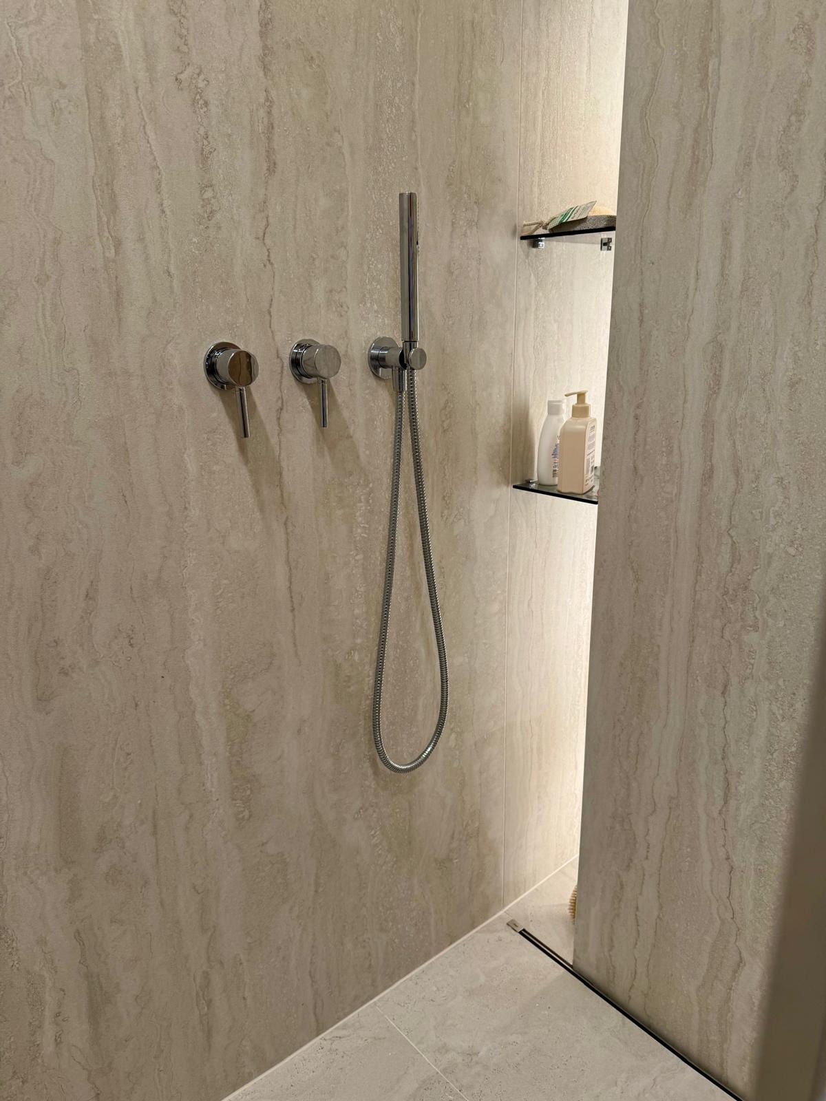 Beżowa łazienka z chromowaną armaturą: bateria, słuchawka prysznicowa i półka z kosmetykami. Minimalistyczny design z subtelnym wzorem na płytkach.