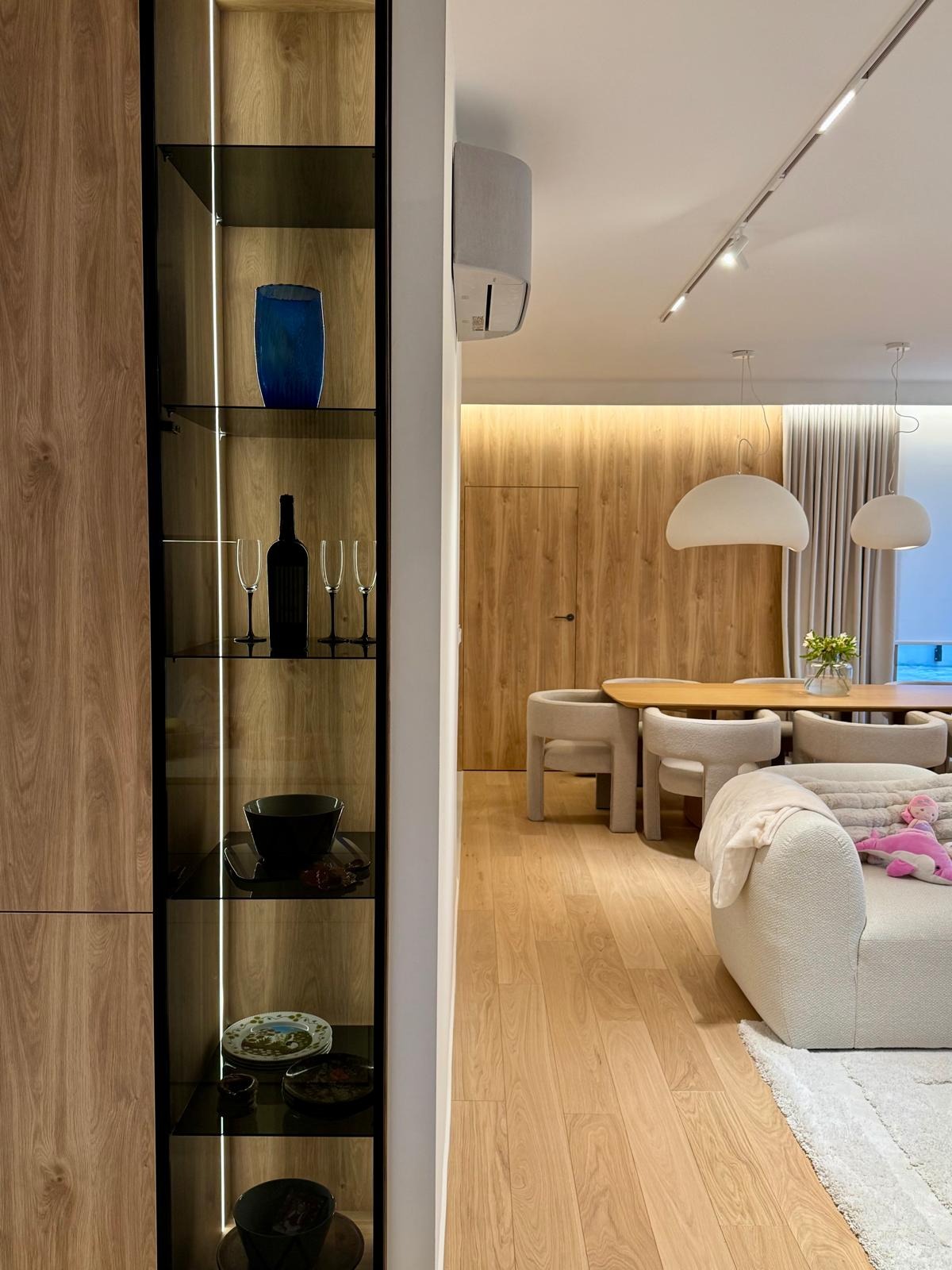 Nowoczesne wnętrze: przeszklona witryna z oświetleniem LED, drewniana ściana z ukrytymi drzwiami, stół z krzesłami i sofa z zabawką, tworzące spójną aranżację.