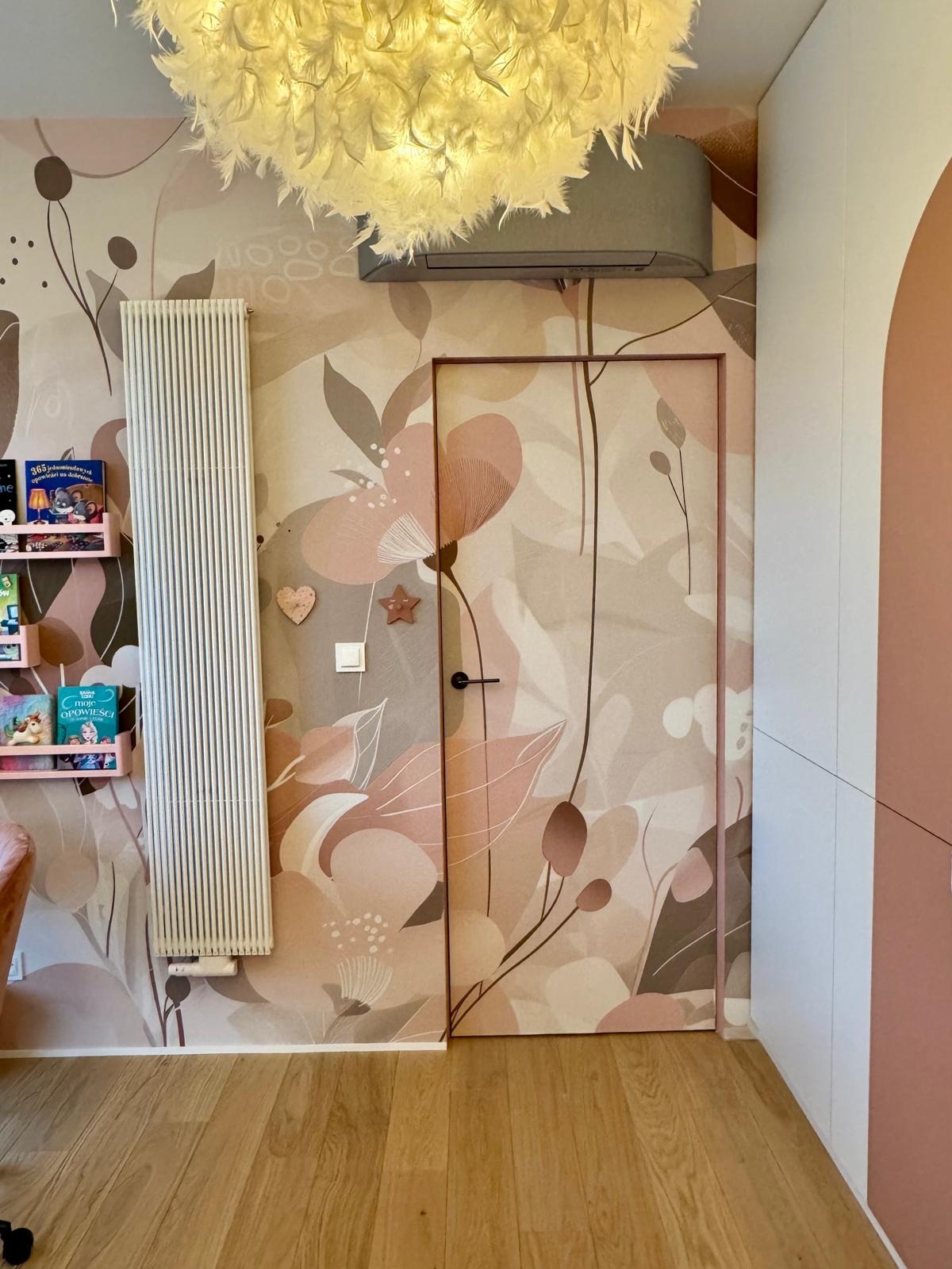 Pokój dziecięcy z tapetą w kwiaty, dopasowaną do drzwi i grzejnika. Pastelowe barwy, drewniana podłoga, półki z książkami i dekoracje na ścianie tworzą spójną całość.