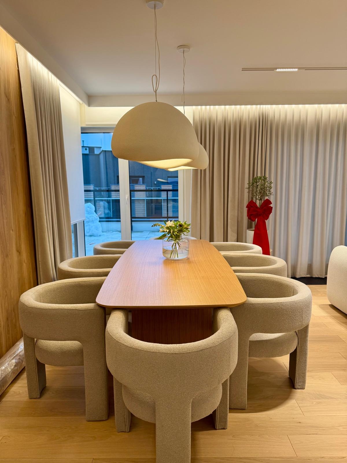 Beżowe krzesła i drewniany stół pod designerską lampą w jasnym wnętrzu. Za oknem widoczny śnieg. Minimalistyczny wystrój z zasłonami i rośliną z czerwoną kokardą.