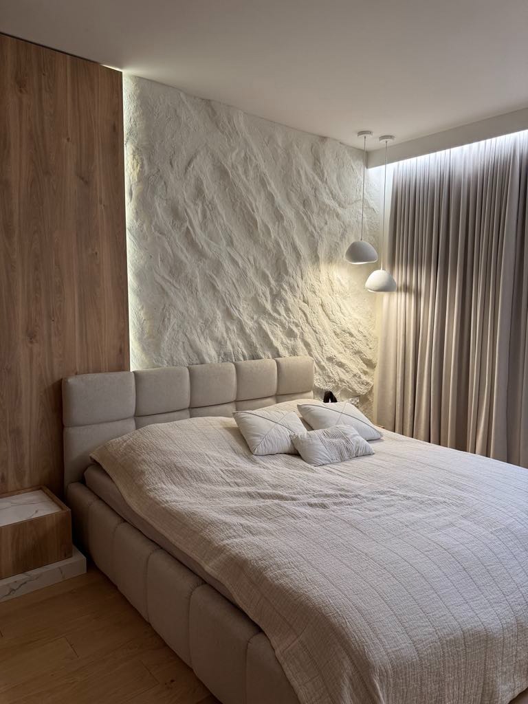Nowoczesna sypialnia z łóżkiem kontynentalnym, drewnianą boazerią i ścianą z fakturą skały, oświetlona wiszącymi lampami. Minimalistyczny design wnętrza.