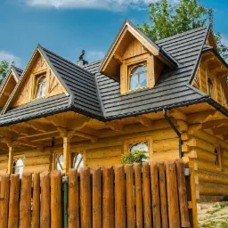 Dom w stylu podhalańskim z drewnianą elewacją i dachem pokrytym gontem blaszanym imitującym drewno, widoczne okna z drewnianymi ramami i drewniany płot.