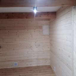 STAN-DACH Stanisław Łuszczek - Wnętrze pomieszczenia wykończonego boazerią drewnianą w dwóch odcieniach, z widoczną instalacją elektryczną, kratką wentylacyjną i drewnianą podłogą.