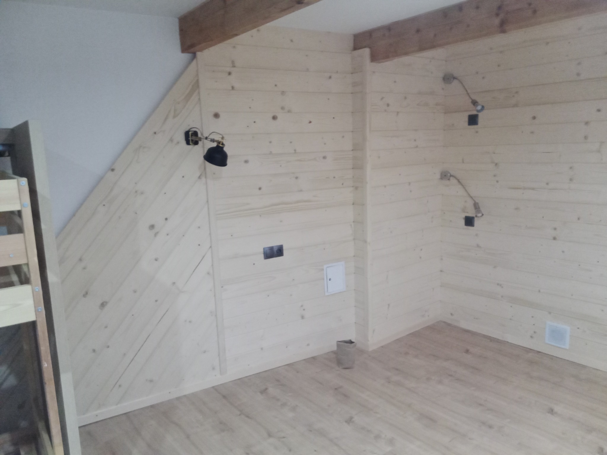 Pomieszczenie wykończone jasnymi panelami drewnianymi na ścianach i podłodze, widoczne punkty oświetleniowe z kablami, otwory na gniazdka elektryczne i kratka wentylacyjna.