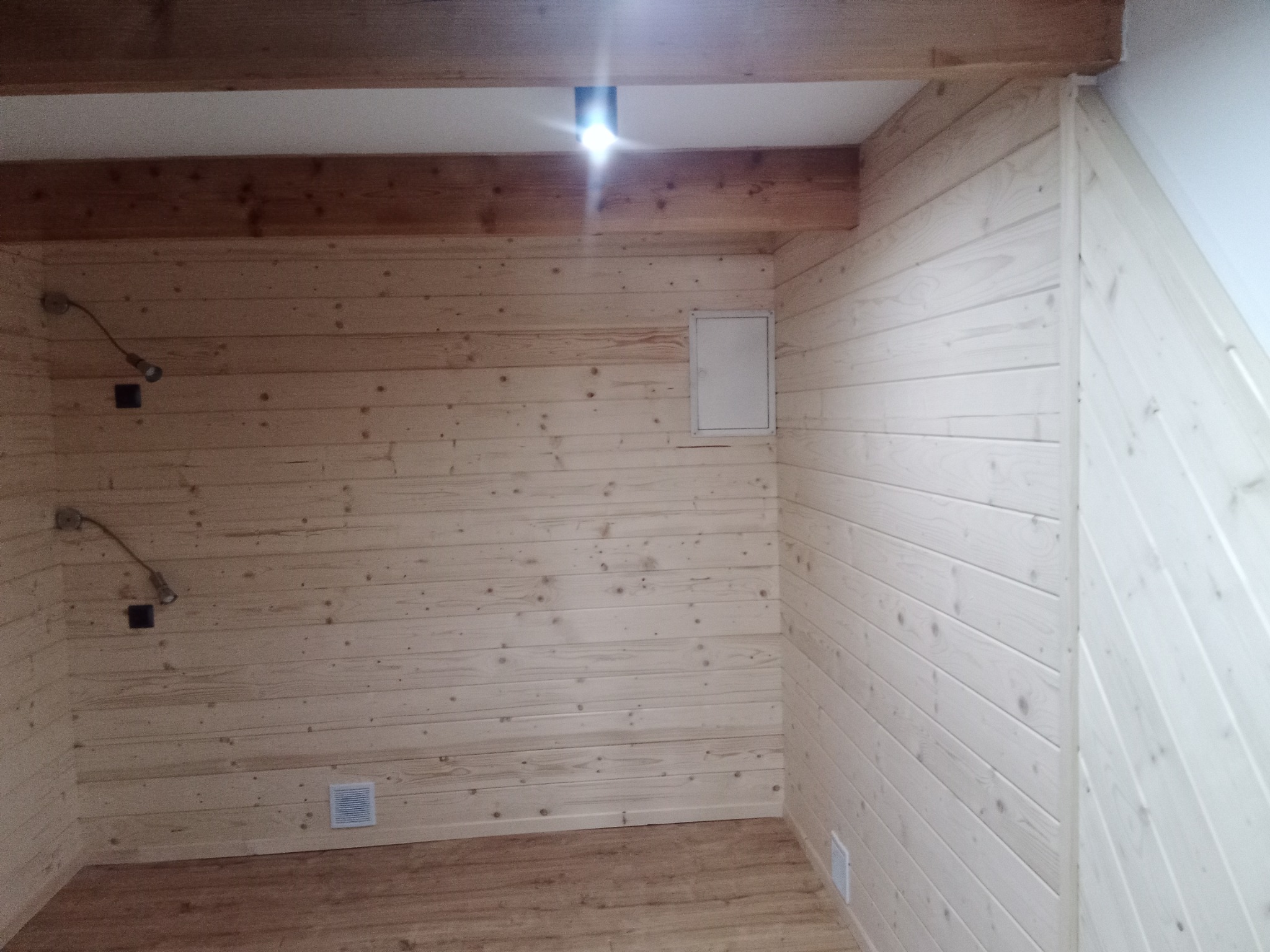 Wnętrze pomieszczenia wykończonego boazerią drewnianą w dwóch odcieniach, z widoczną instalacją elektryczną, kratką wentylacyjną i drewnianą podłogą.