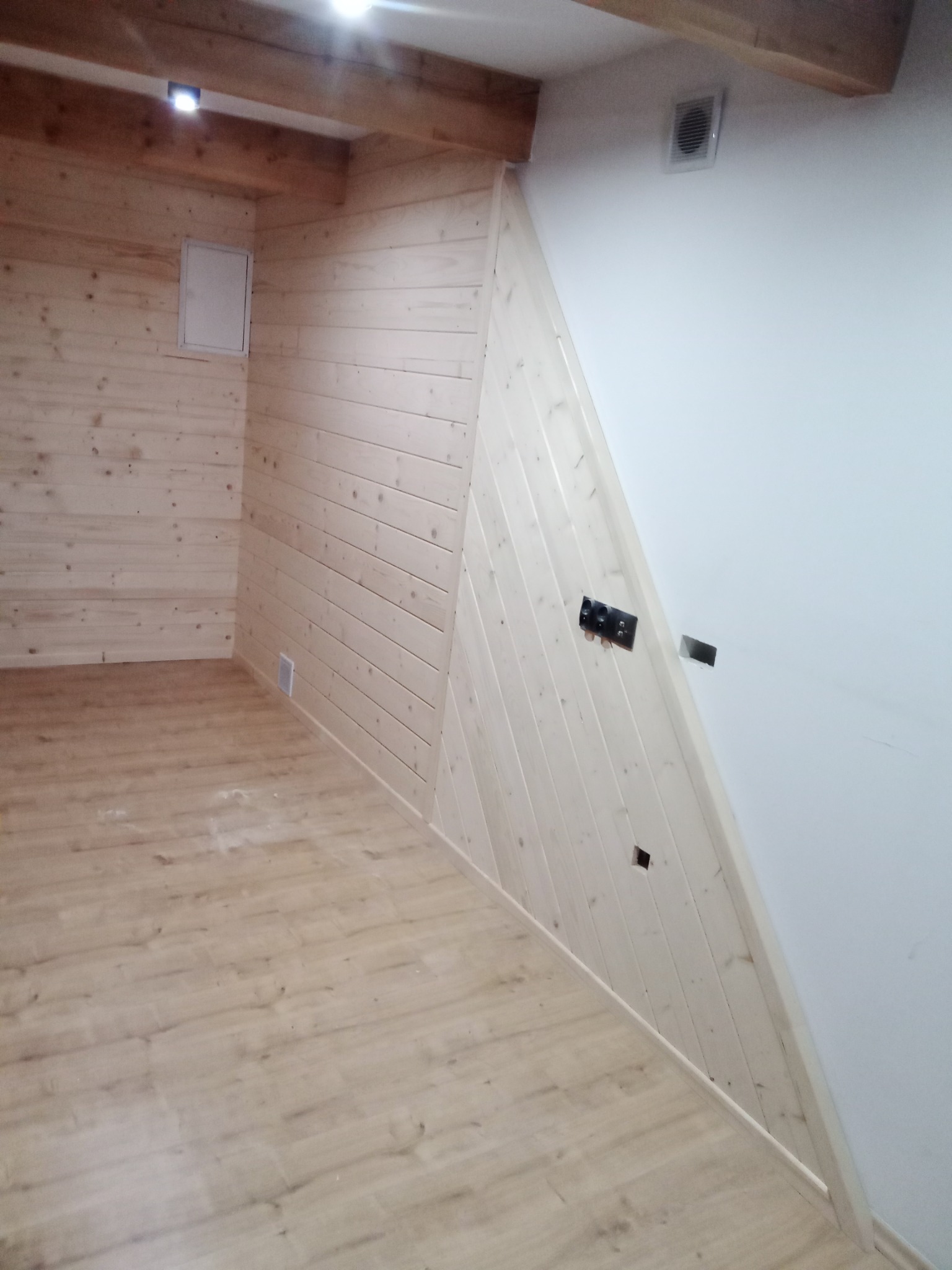 Wnętrze pokoju z drewnianą boazerią na ścianach, drewnianymi belkami stropowymi, panelami podłogowymi i częściowo wykończoną ścianą z gipsu z puszkami elektrycznymi.