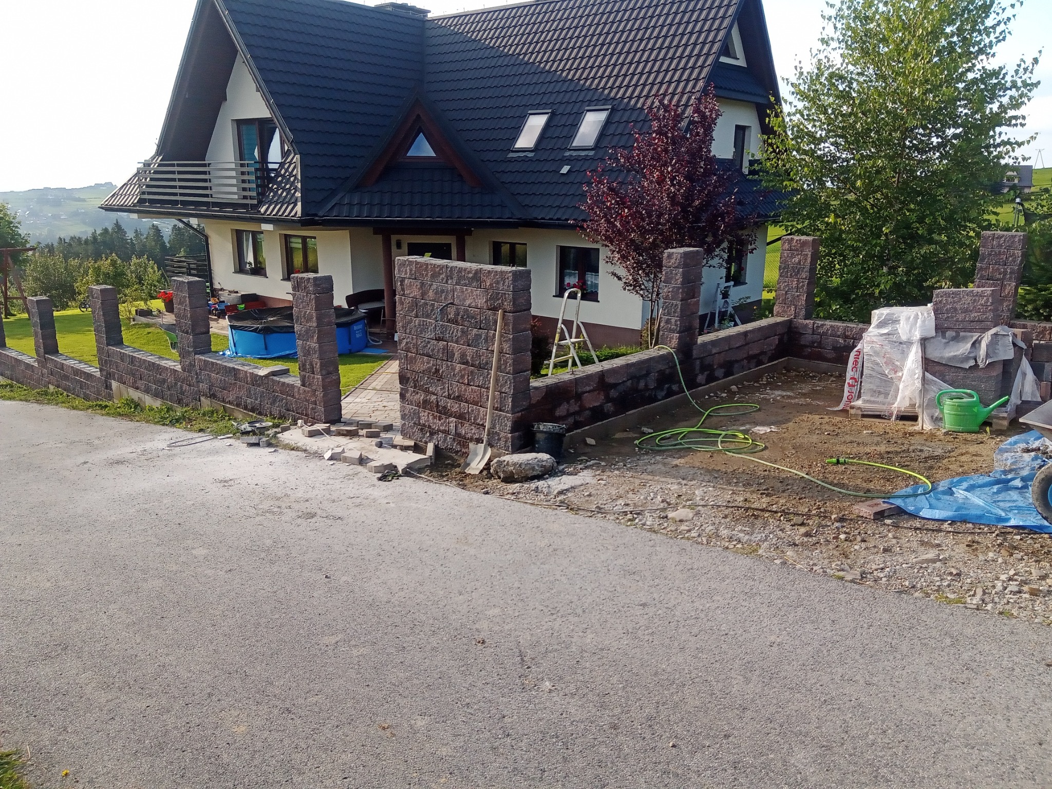 Budowa ogrodzenia z bloczków imitujących kamień, widoczny fragment posesji z domem i ogrodem, narzędzia budowlane i materiały przygotowane do pracy.