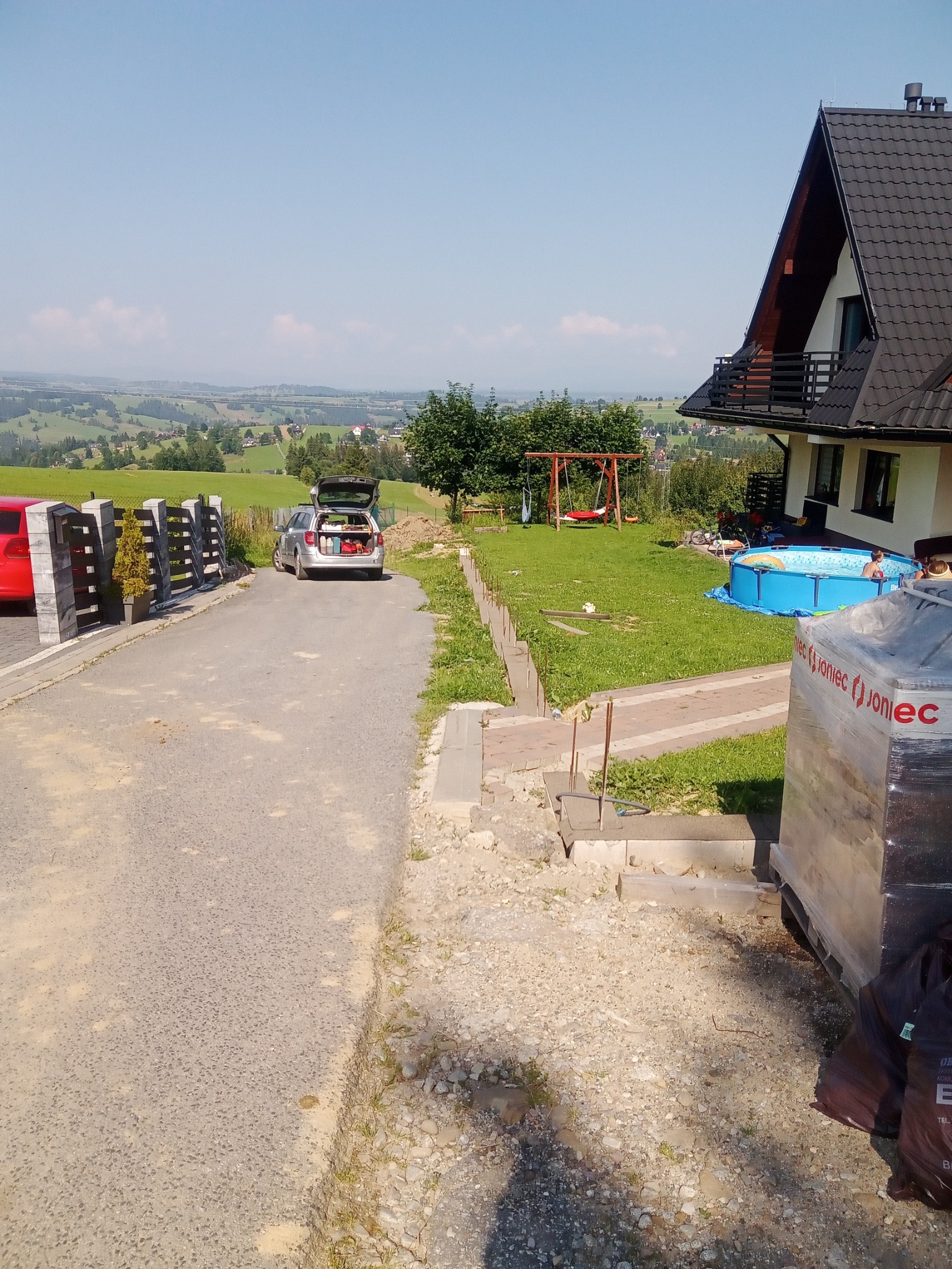 Budowa ogrodzenia panelowego: widok na posesję z nieukończonym ogrodzeniem z betonowych słupków, zaparkowany samochód z otwartym bagażnikiem, dom z ciemnym dachem, basen na trawniku i malowniczy...