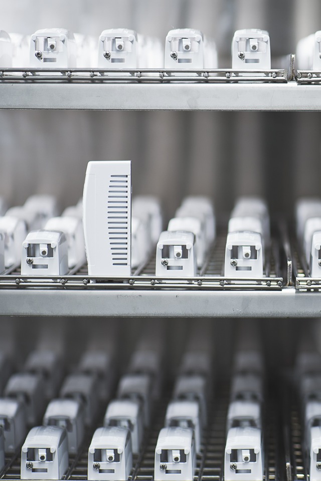 Białe elementy systemu wentylacyjnego na metalowych półkach w sterylnym pomieszczeniu, jeden element pionowy z widocznymi żebrami wentylacyjnymi.