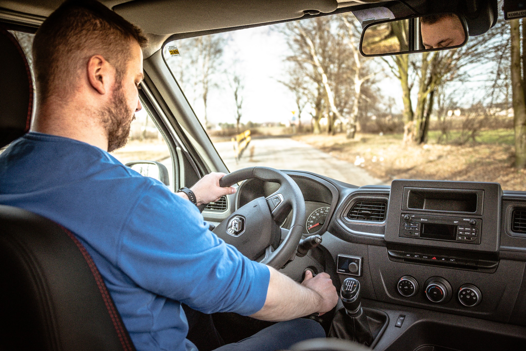 Kierowca w niebieskiej koszuli za kierownicą busa, widok na drogę przez przednią szybę, drzewa w tle.