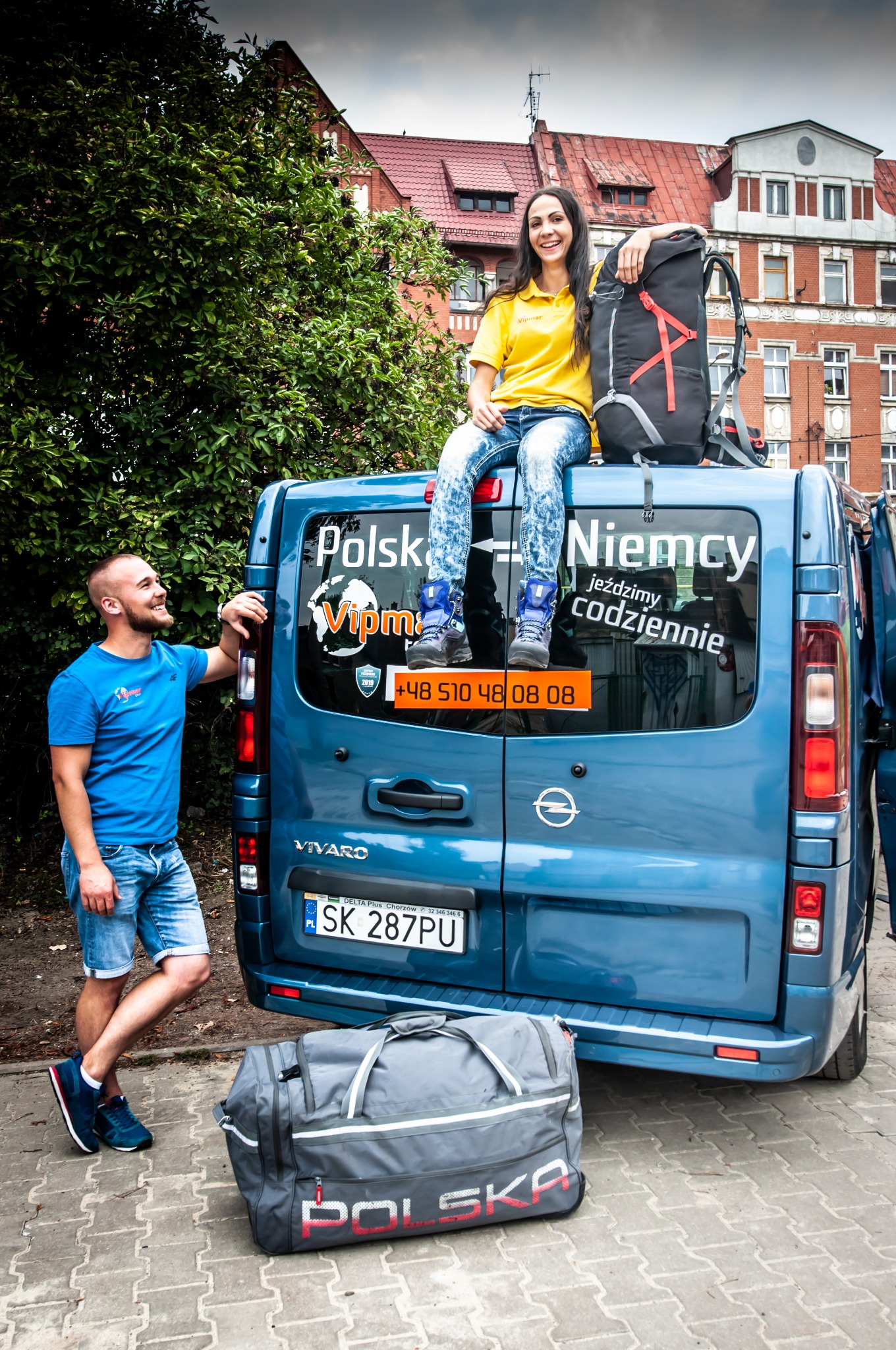 Uśmiechnięta kobieta siedzi na dachu niebieskiego busa z naklejkami 'Polska Niemcy jeździmy codziennie', obok stoi mężczyzna, a na ziemi leży duża torba podróżna.