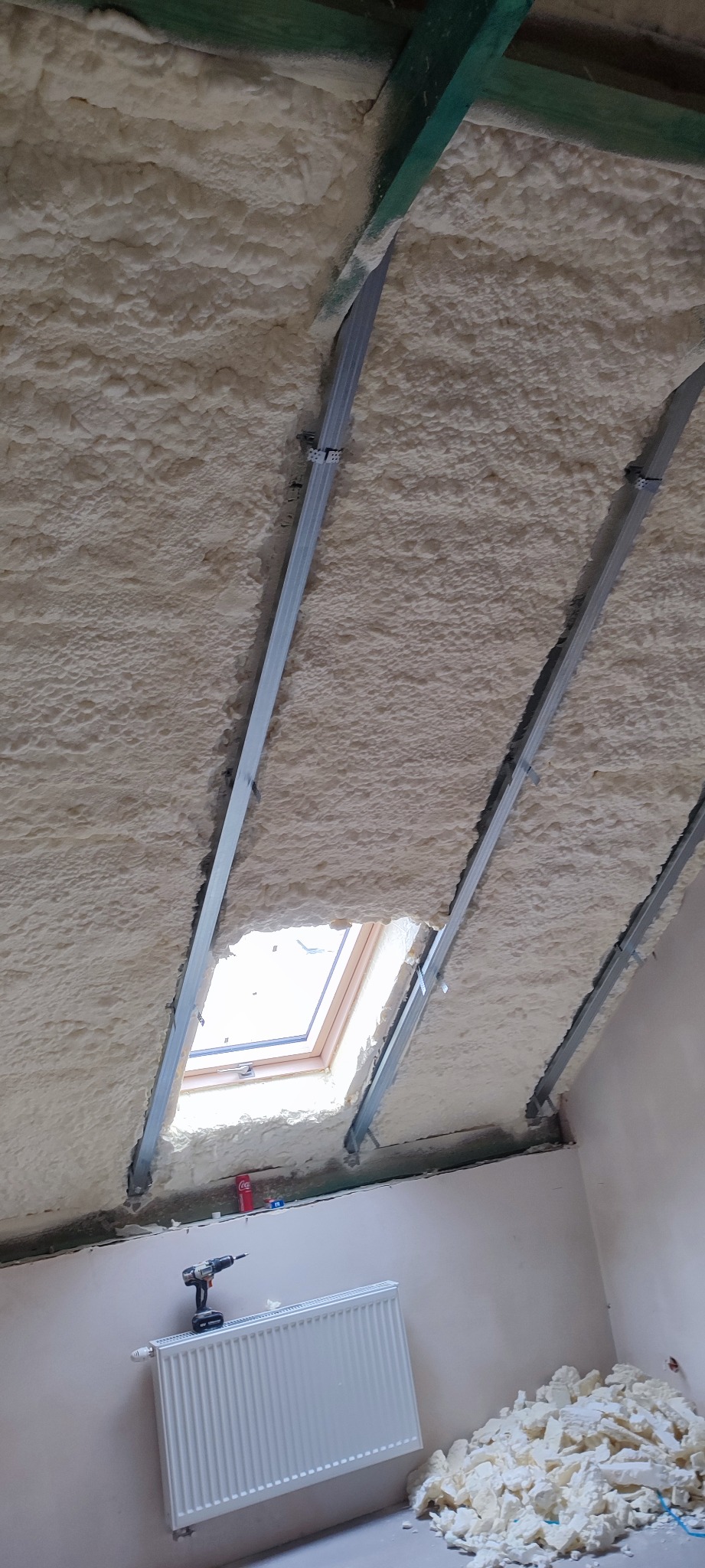 Poddasze z oknem dachowym, ocieplone pianką poliuretanową, widoczne metalowe profile konstrukcyjne, grzejnik i wiertarka akumulatorowa na ścianie.