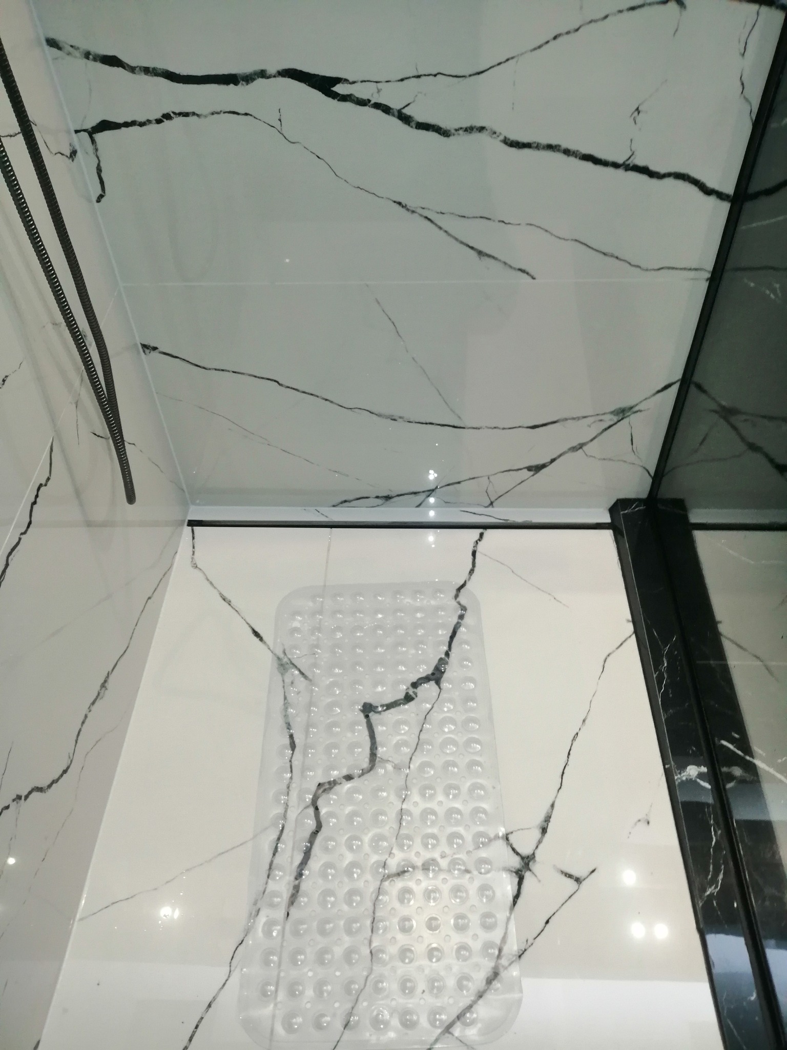 Kabina prysznicowa wyłożona białymi płytkami imitującymi marmur z czarnymi żyłami, z antypoślizgową, przezroczystą matą na podłodze.