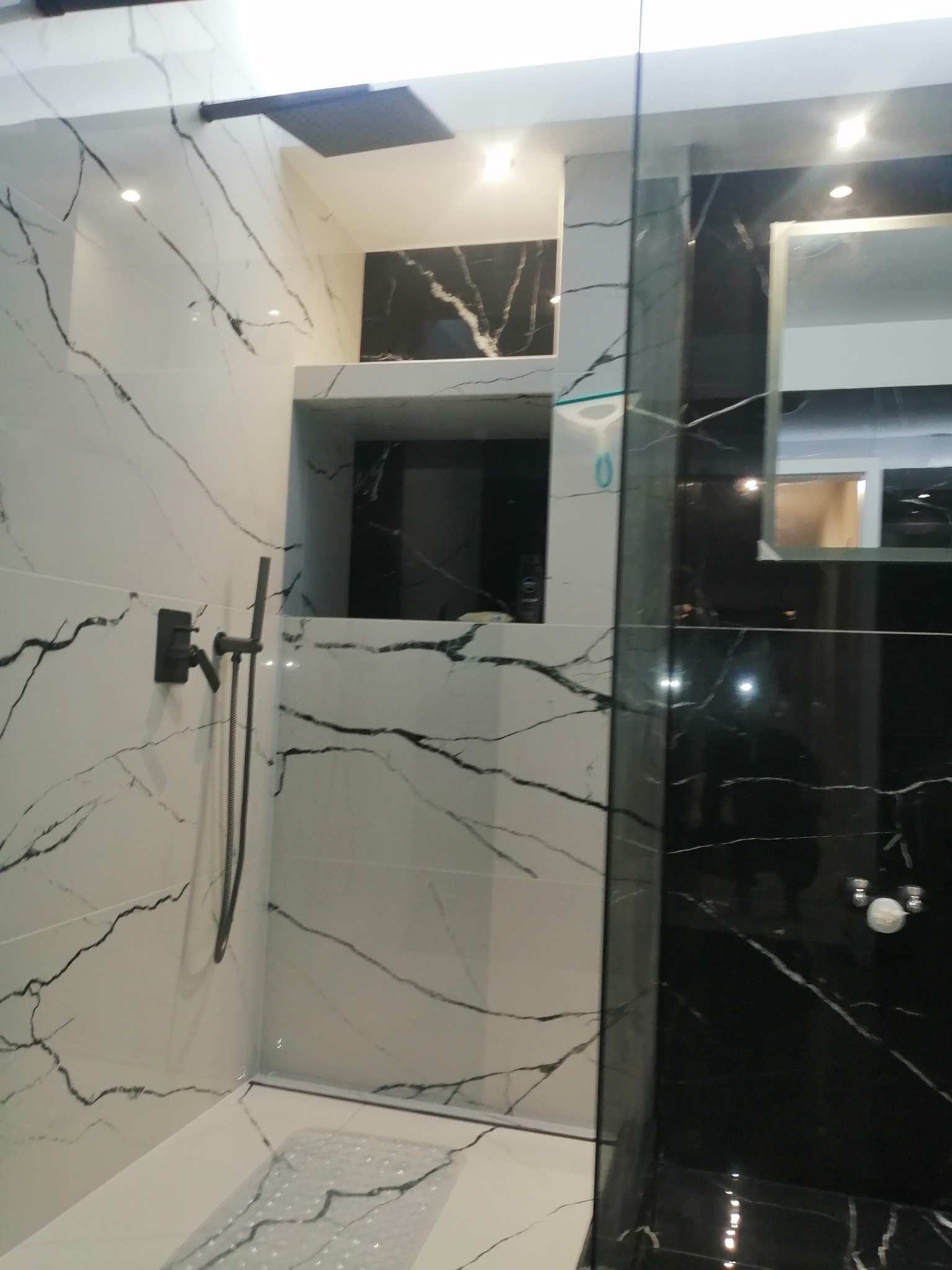 Nowoczesna kabina prysznicowa z czarno-białymi płytkami imitującymi marmur, czarny zestaw prysznicowy, wnęka w ścianie wyłożona czarnym marmurem, widoczny fragment lustra.