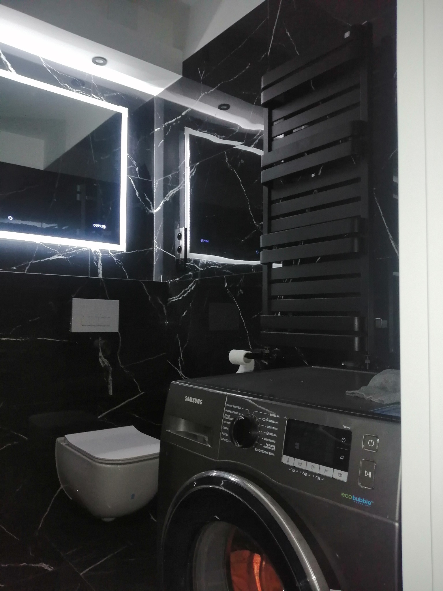 Nowoczesna łazienka w Warszawie z czarnymi marmurowymi płytkami, podświetlanymi lustrami, czarnym grzejnikiem drabinkowym i pralką Samsung. Uwagę zwraca nietypowe umiejscowienie toalety obok pralki.
