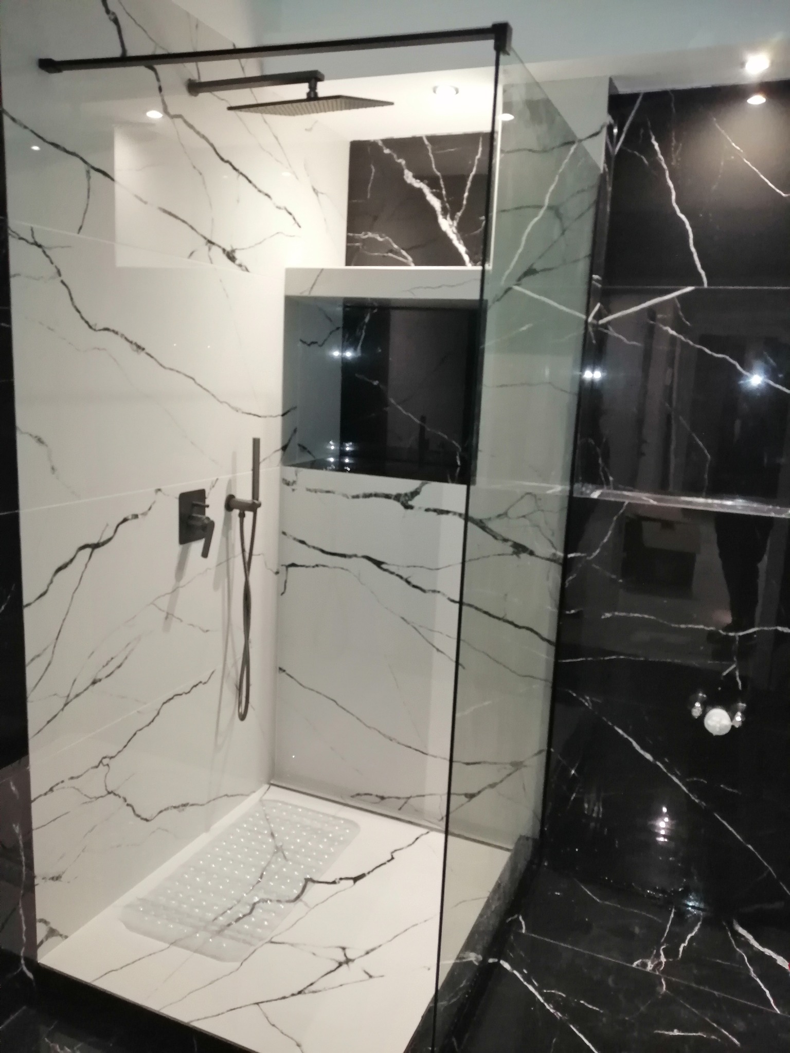 Nowoczesna kabina prysznicowa walk-in z czarno-białymi płytkami imitującymi marmur, czarna armatura i odpływ liniowy, oświetlenie punktowe.