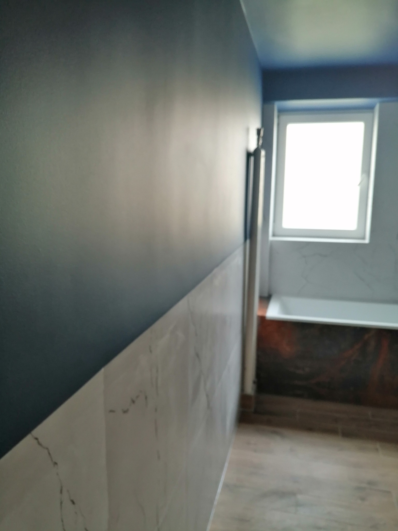 Ściana w łazience z pomalowaną górną częścią na ciemny niebieski kolor i dolną wyłożoną białymi płytkami imitującymi marmur, widoczna wanna i fragment podłogi z paneli.