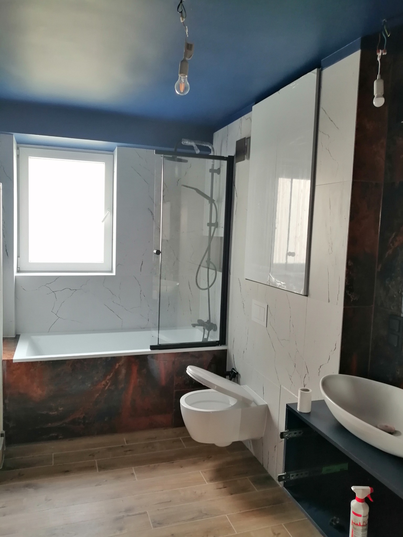 Nowoczesna łazienka z marmurowymi płytkami, drewnopodobną podłogą, wiszącą toaletą i umywalką nablatową, niebieski sufit z wiszącą żarówką.