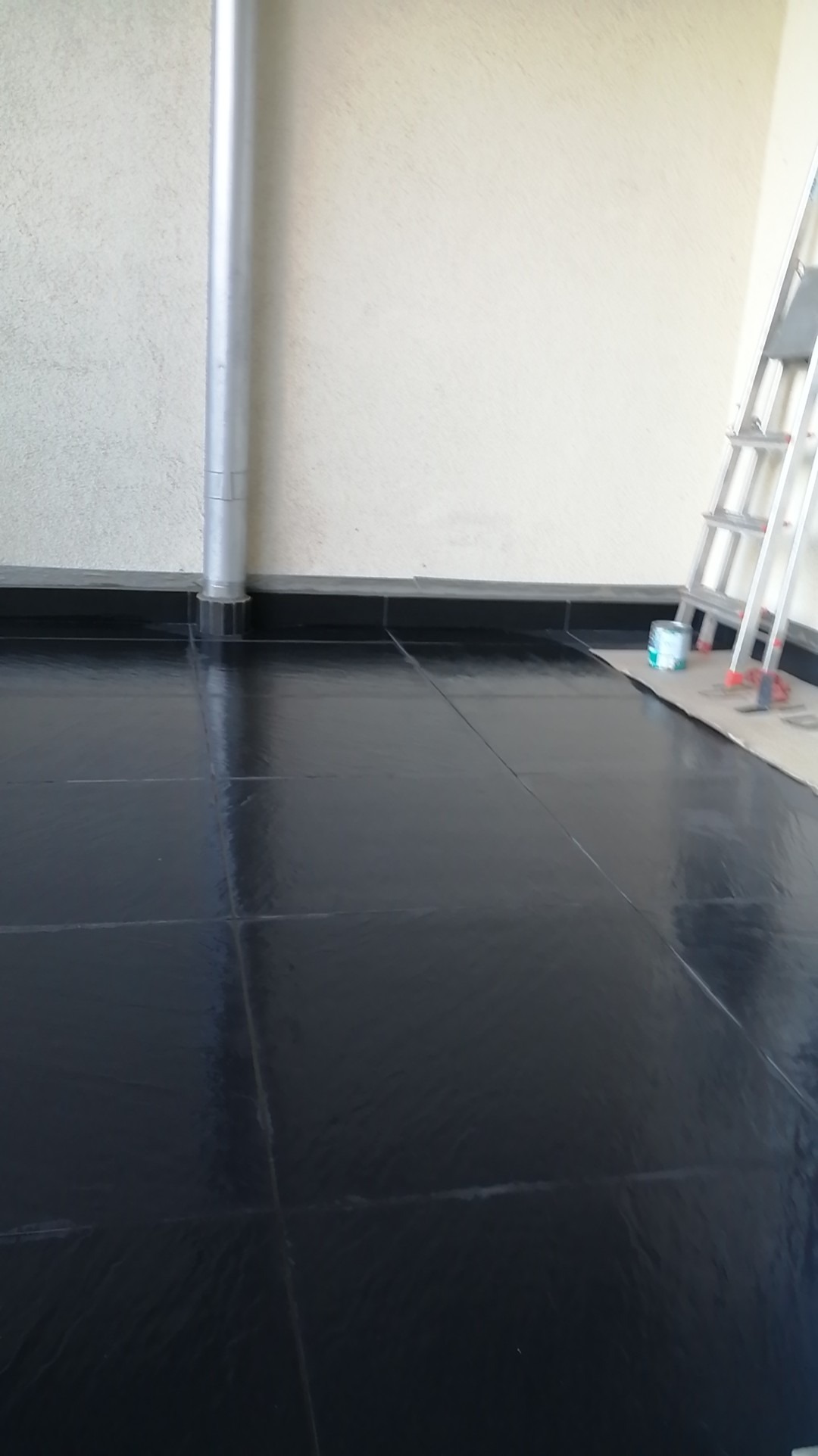 Błyszcząca, czarna podłoga wyłożona kwadratowymi płytkami, z widoczną metalową rurą pionową przy ścianie i drabiną opartą o ścianę w tle, sugerująca prace wykończeniowe w pomieszczeniu.