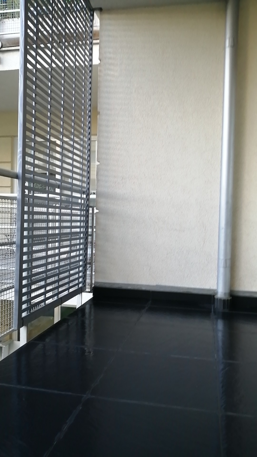 Balkon z czarną, błyszczącą podłogą z płytek, szarą metalową balustradą z poziomymi szczeblami i roletą w tle.