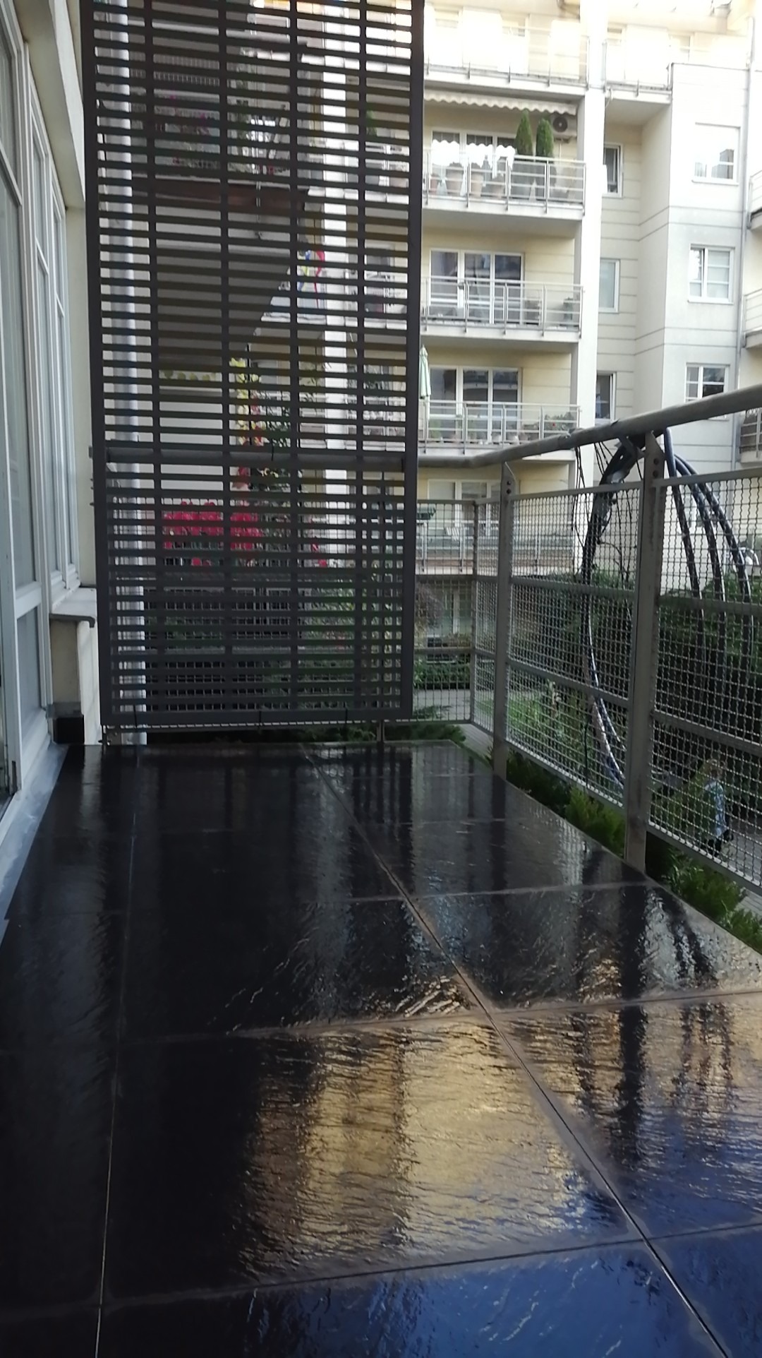 Błyszcząca, ciemna podłoga balkonu z płytkami imitującymi kamień, widok na metalową kratę i budynek mieszkalny w tle.