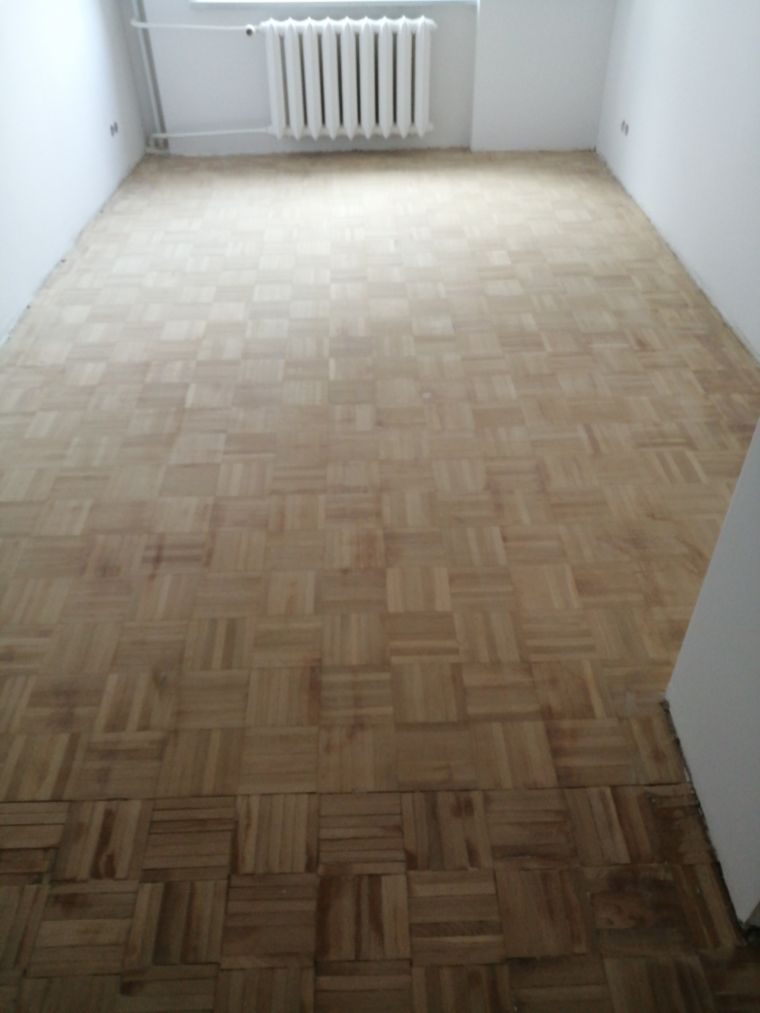 Parkiet ułożony we wzór szachownicy w pustym pokoju z białymi ścianami i grzejnikiem pod oknem.