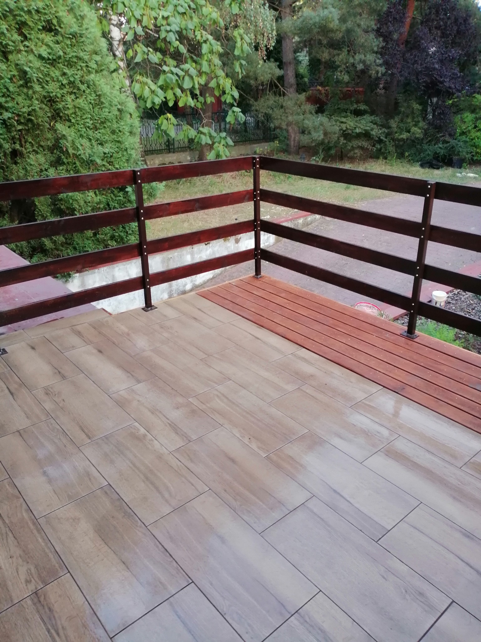 Wyłożony płytkami taras z drewnianą balustradą, widok na zieleń ogrodu w tle.