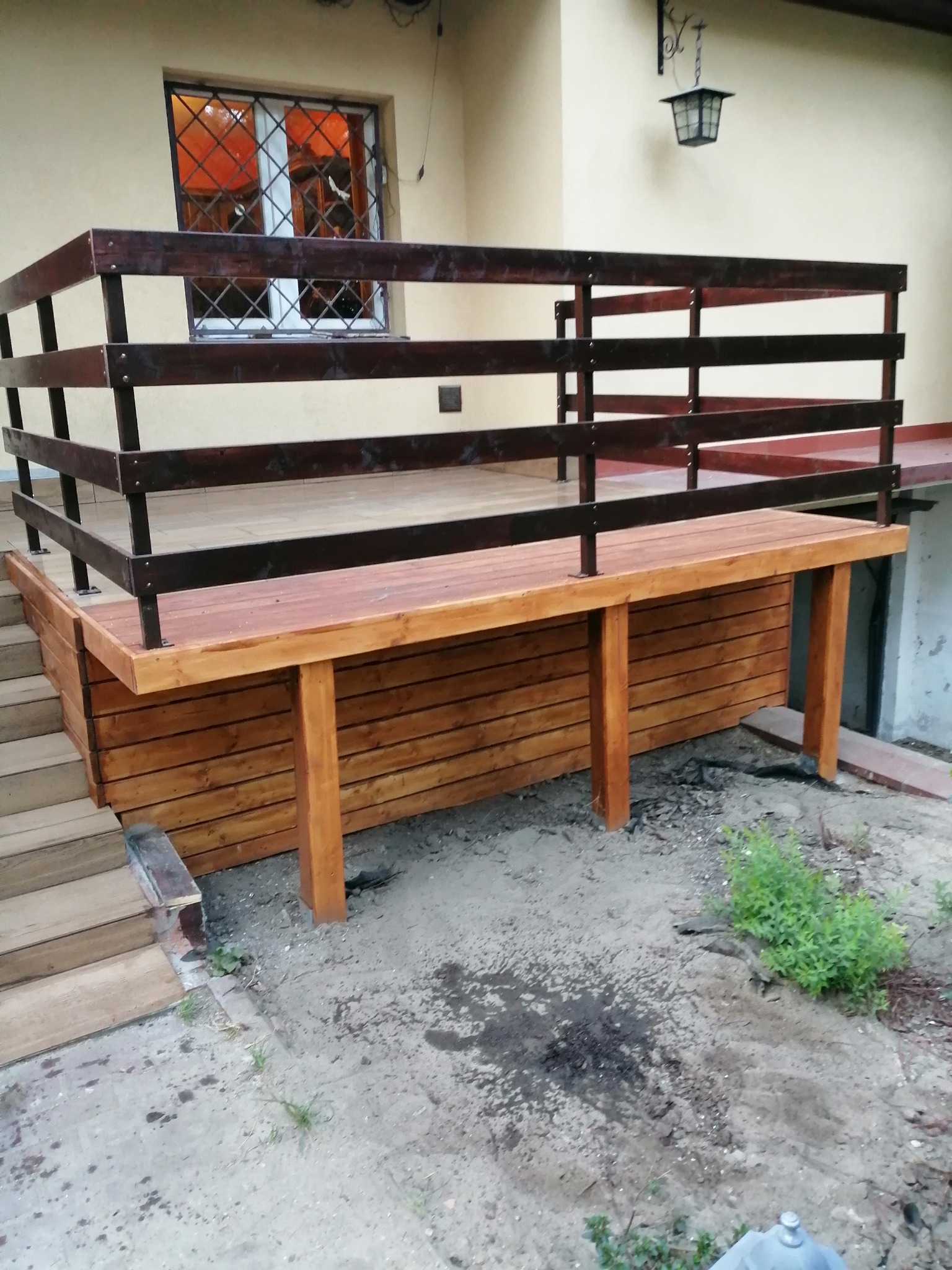 Drewniany taras z brązową balustradą i schodami, przylegający do beżowego budynku z oknem z kratami i wiszącą latarnią, widok z poziomu gruntu.