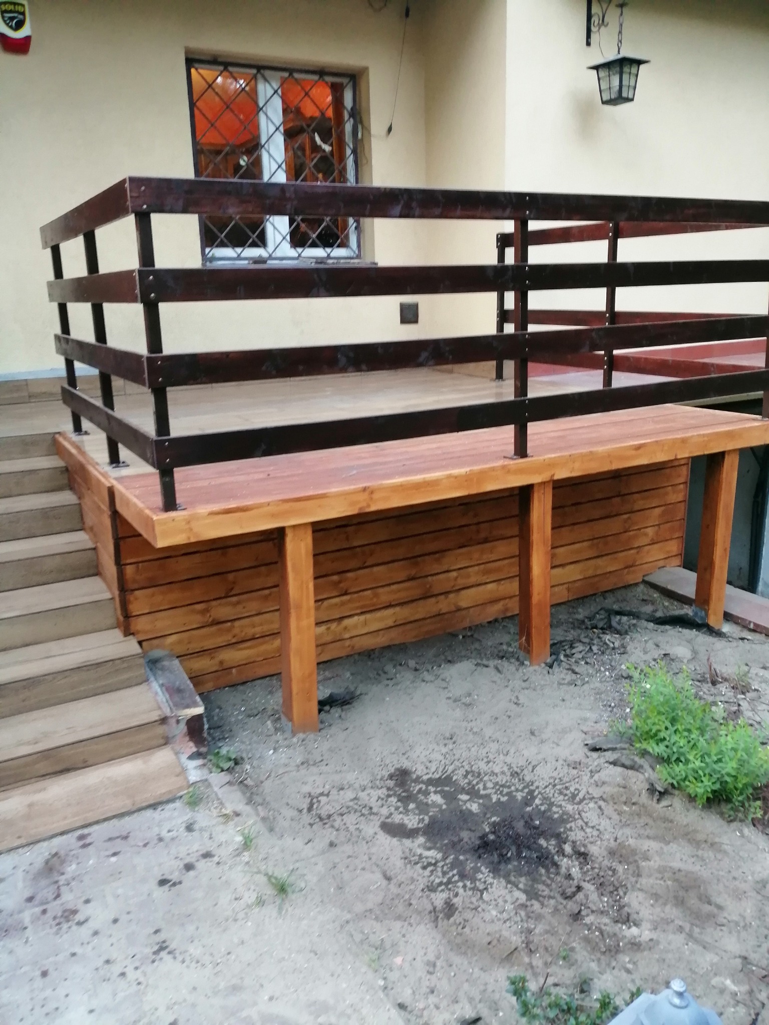 Nowo wybudowany drewniany taras z brązową balustradą, przylegający do domu, z widocznymi schodami wejściowymi i lampą wiszącą nad tarasem.