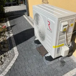 Biała jednostka zewnętrzna pompy ciepła marki Rotenso, zamontowana na betonowych podstawach obok ścieżki z szarej kostki brukowej, na tle żółtej ściany budynku z ozdobami okiennymi.