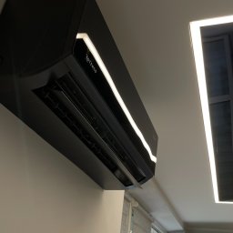 TRAVEL INVEST GROUP SPÓŁKA Z OGRANICZONĄ ODPOWIEDZIALNOŚCIĄ - Czarna, nowoczesna klimatyzacja Rotenso z podświetleniem LED zamontowana na ścianie w jasnym pomieszczeniu z oknem.