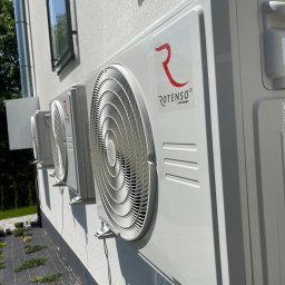 TRAVEL INVEST GROUP SPÓŁKA Z OGRANICZONĄ ODPOWIEDZIALNOŚCIĄ - Trzy jednostki zewnętrzne klimatyzacji marki Rotenso zamontowane na elewacji budynku z widocznym oknem i zagospodarowanym terenem wokół.