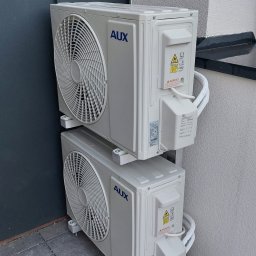 TRAVEL INVEST GROUP SPÓŁKA Z OGRANICZONĄ ODPOWIEDZIALNOŚCIĄ - Dwa klimatyzatory AUX zamontowane jeden nad drugim na zewnątrz budynku, widoczne przewody i naklejki ostrzegawcze.