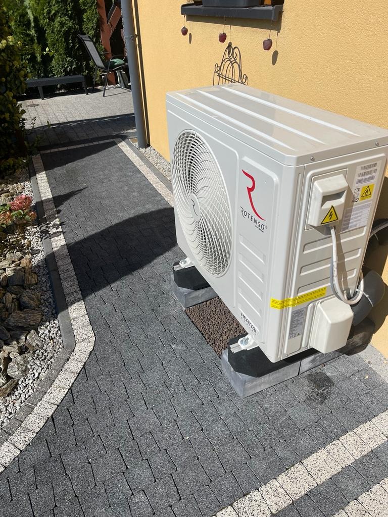 Biała jednostka zewnętrzna pompy ciepła marki Rotenso, zamontowana na betonowych podstawach obok ścieżki z szarej kostki brukowej, na tle żółtej ściany budynku z ozdobami okiennymi.