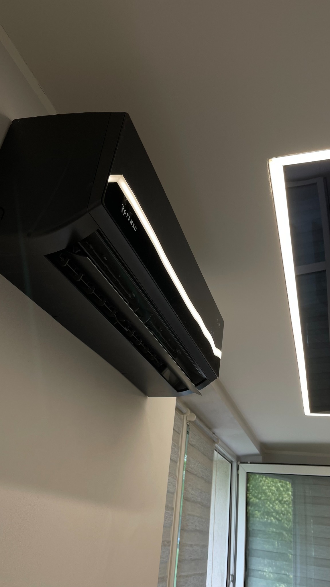 Czarna, nowoczesna klimatyzacja Rotenso z podświetleniem LED zamontowana na ścianie w jasnym pomieszczeniu z oknem.