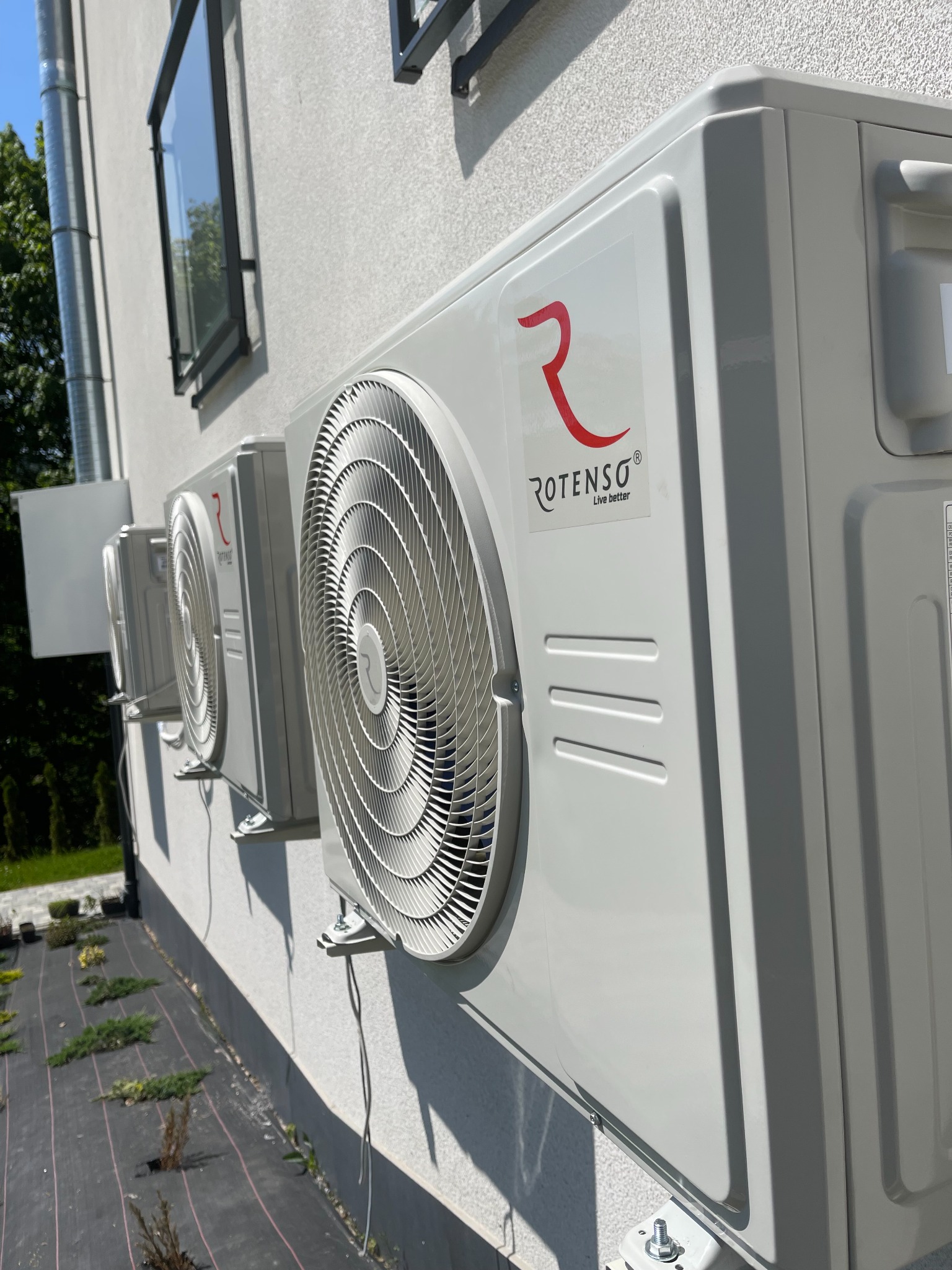 Trzy jednostki zewnętrzne klimatyzacji marki Rotenso zamontowane na elewacji budynku z widocznym oknem i zagospodarowanym terenem wokół.