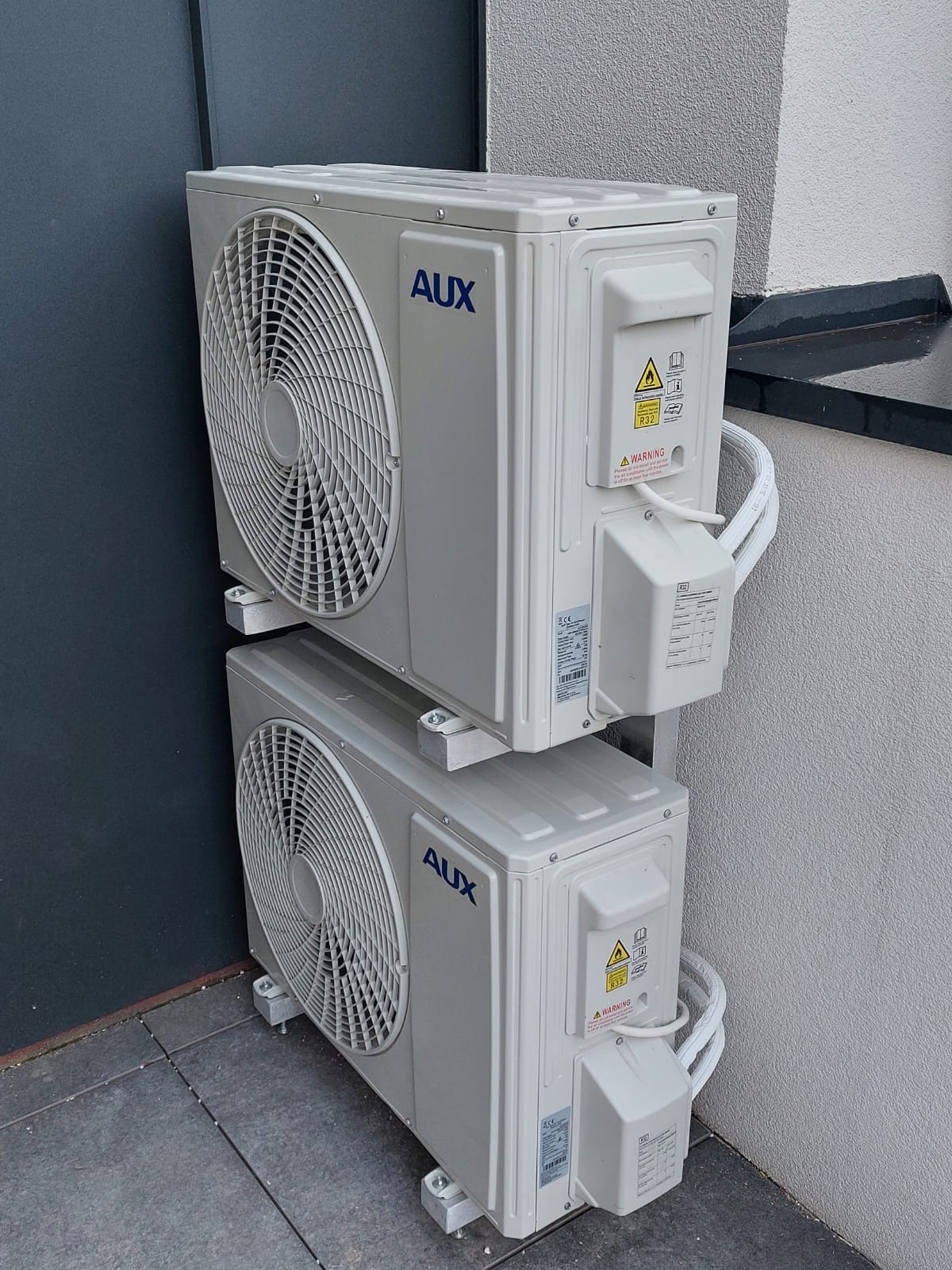 Dwa klimatyzatory AUX zamontowane jeden nad drugim na zewnątrz budynku, widoczne przewody i naklejki ostrzegawcze.