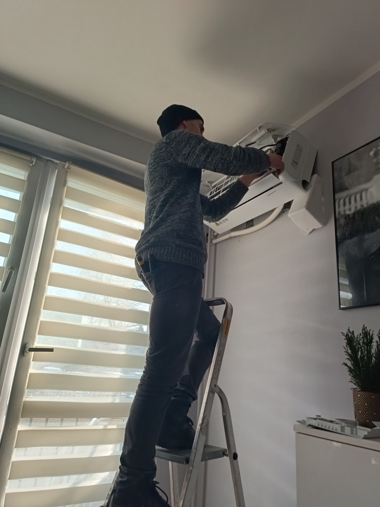 Mężczyzna w czapce, stojący na aluminiowej drabinie, naprawia białą klimatyzację ścienną w pomieszczeniu z roletami okiennymi i dekoracjami.