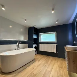Nowoczesna łazienka z wolnostojącą wanną, drewnopodobną podłogą, umywalką nablatową z podświetlanym lustrem, grzejnikiem panelowym i pralką ukrytą w zabudowie.