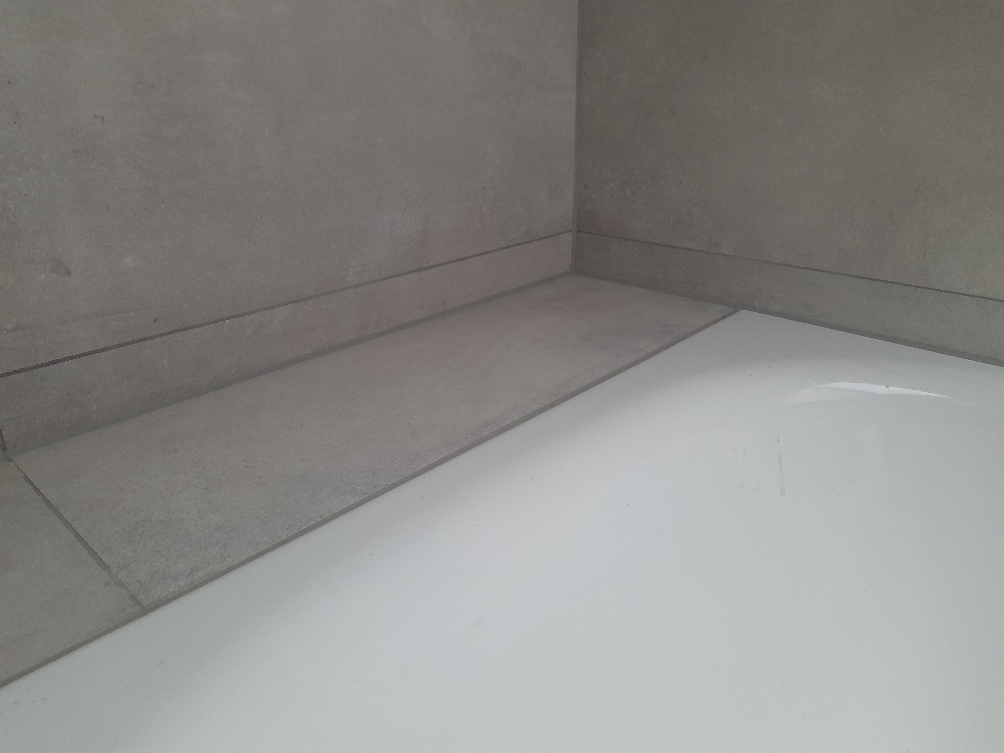 Szare płytki ceramiczne na ścianach i podłodze w rogu łazienki, widoczna biała wanna lub brodzik.