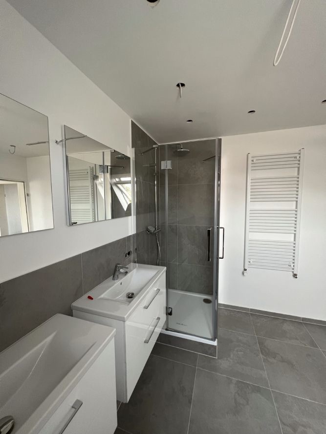 Nowoczesna łazienka z szarymi płytkami, kabiną prysznicową typu walk-in, białą szafką z umywalką i grzejnikiem drabinkowym, widoczne niedokończone elementy oświetlenia sufitowego.