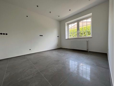 Pusty pokój z szarymi płytkami podłogowymi, białymi ścianami z przygotowanymi punktami elektrycznymi, białym oknem z widokiem na zieleń i białym grzejnikiem pod oknem.