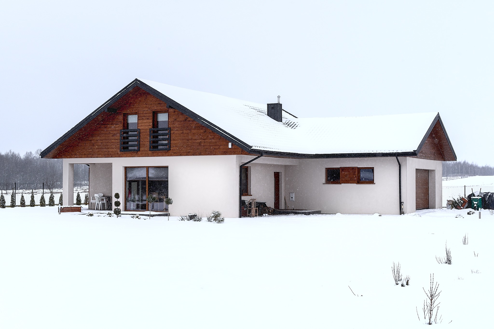 Zimowy krajobraz z białym, parterowym domem z drewnianą elewacją pod dachem, pokrytym śniegiem, widoczne zaspy śnieżne i lekko ośnieżone drzewa w tle.