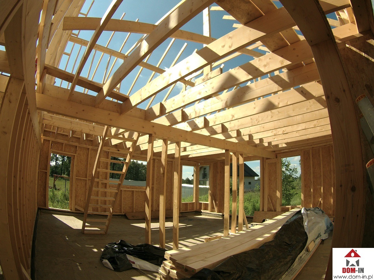 Wnętrze domu w trakcie budowy z drewnianą konstrukcją szkieletową, widoczne belki stropowe, ściany z płyt OSB i drabina wewnątrz. Widok na zewnątrz przez otwory okienne, częściowo pokryte folią...