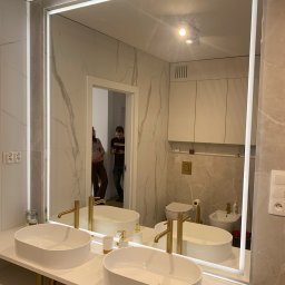 FUNKCJA. Studio wnętrz Aleksandra Chruściel-Bujnowska - Nowoczesna łazienka z podwójną umywalką nablatową, złotą armaturą i lustrem LED. W tle widoczna toaleta i bidet w jasnej tonacji.