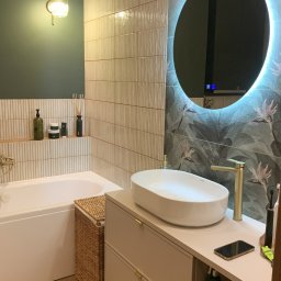 FUNKCJA. Studio wnętrz Aleksandra Chruściel-Bujnowska - Nowoczesna łazienka z okrągłym lustrem LED, beżową szafką z umywalką i drewnianą podłogą. Na ścianie płytki w odcieniach beżu i tapeta z motywem roślinnym.