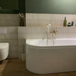 FUNKCJA. Studio wnętrz Aleksandra Chruściel-Bujnowska - Elegancka łazienka z białą wanną, złotą armaturą i beżowymi płytkami. Na półce dekoracje i kosmetyki. Ciemnozielona ściana nadaje wnętrzu nowoczesny charakter.