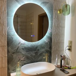 FUNKCJA. Studio wnętrz Aleksandra Chruściel-Bujnowska - Nowoczesna łazienka z okrągłym lustrem LED, umywalką nablatową i zielonymi akcentami. Widoczne płytki z motywem roślinnym i elegancka lampa ścienna.