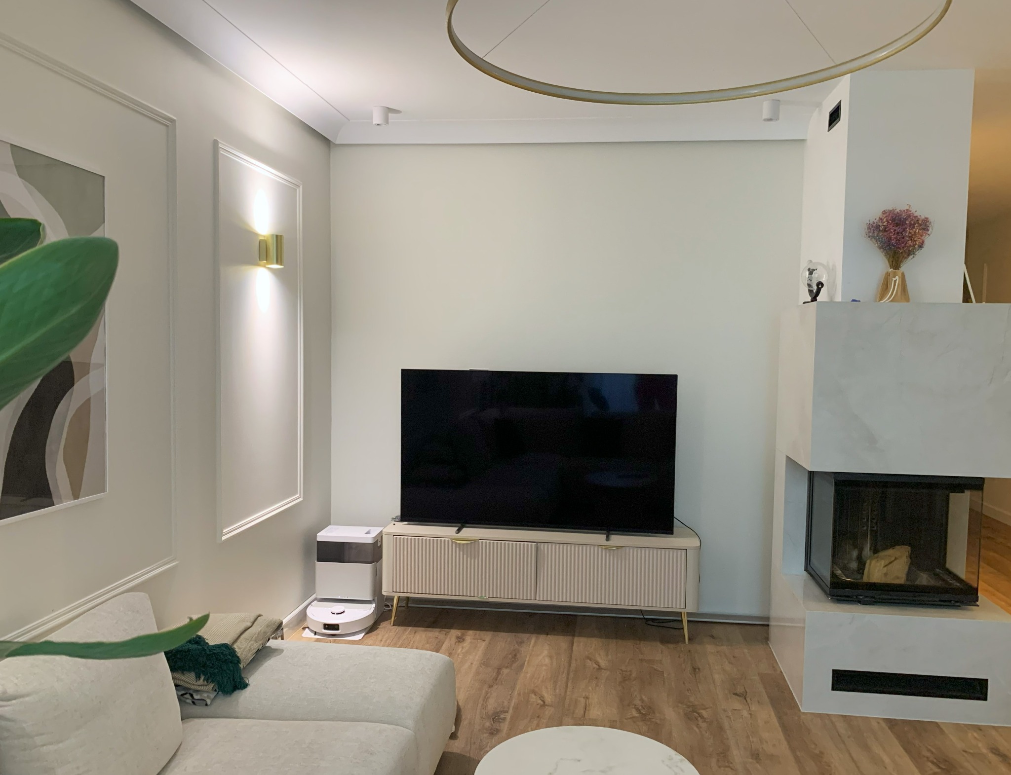 Jasne wnętrze salonu z kominkiem obok telewizora, robot sprzątający przy sofie, minimalistyczne oświetlenie. Ściany zdobią sztukaterie i nowoczesny obraz.